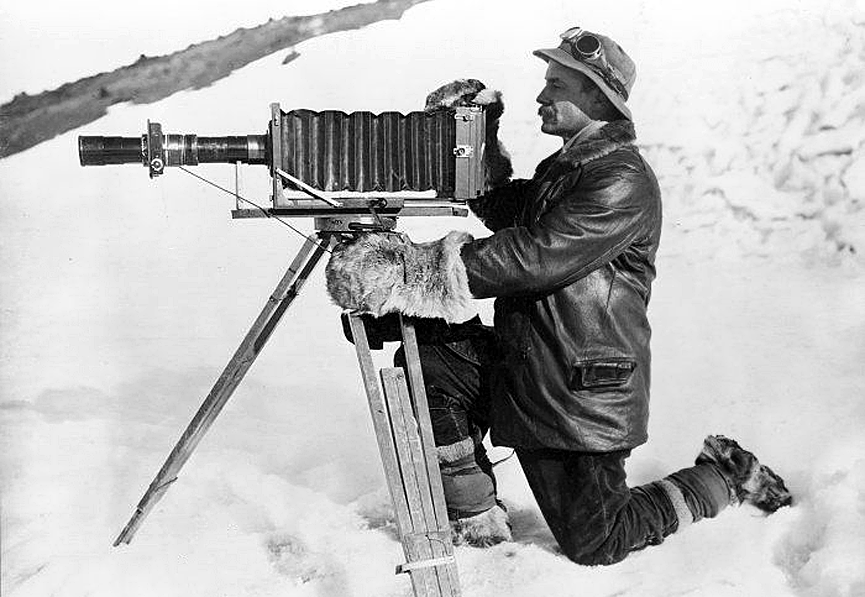 Herbert Ponting se arrodilla en la nieve mirando a través de su cámara montada en un trípode, en enero de 1912. Ponting Collection
